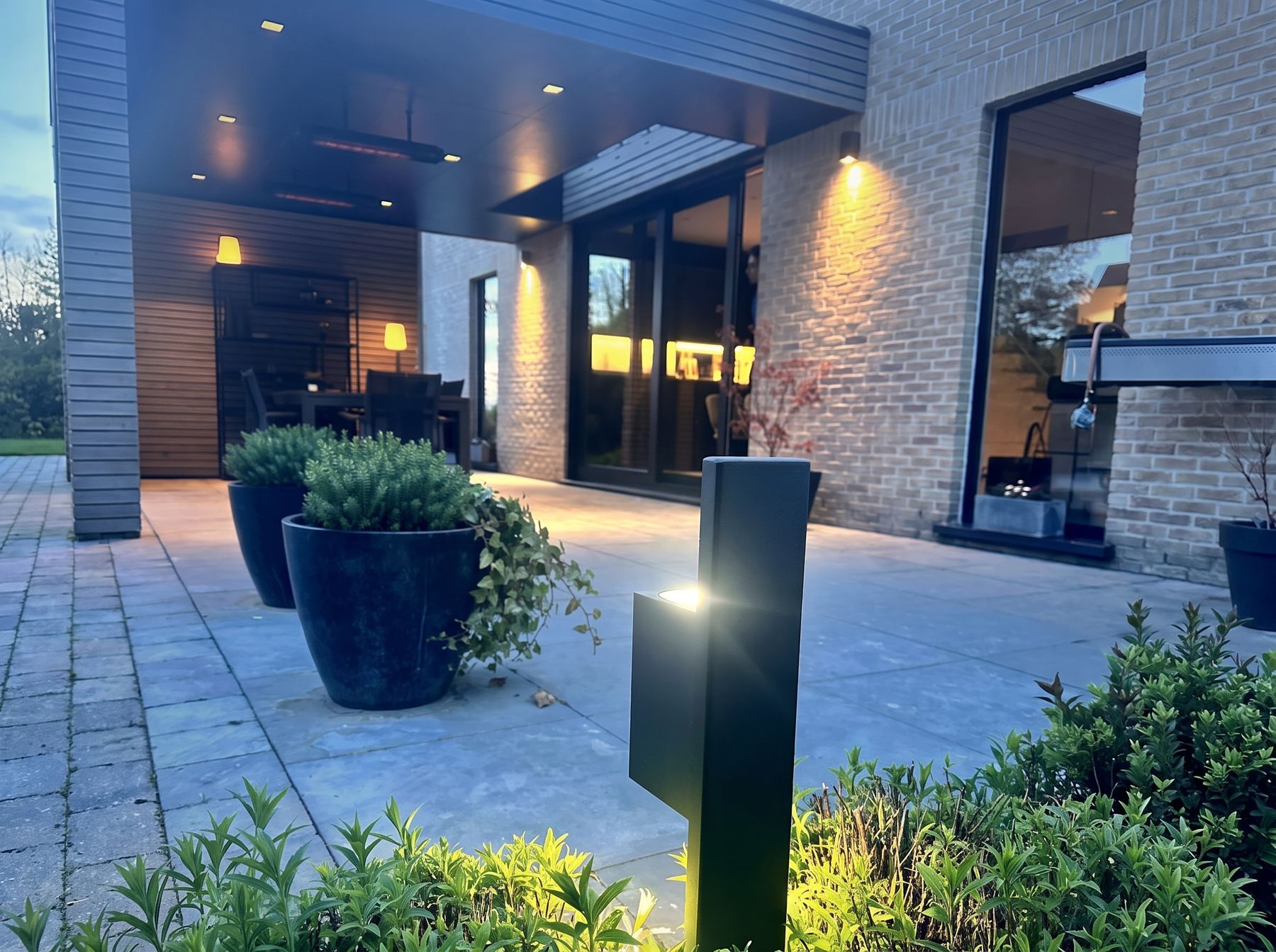 Mise en situation (décor créé par IA) : borne lumineuse en acier noir éclairant une terrasse au crépuscule