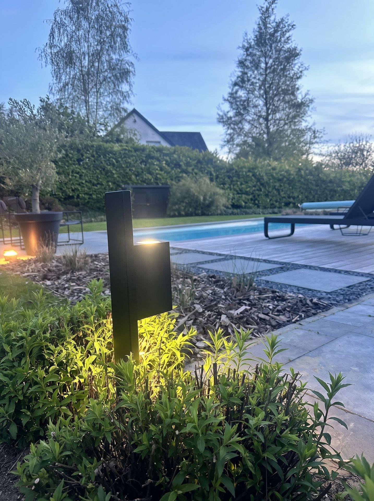 Mise en situation (décor créé par IA) : borne lumineuse en acier noir allumée au crépuscule, dans un jardin avec piscine
