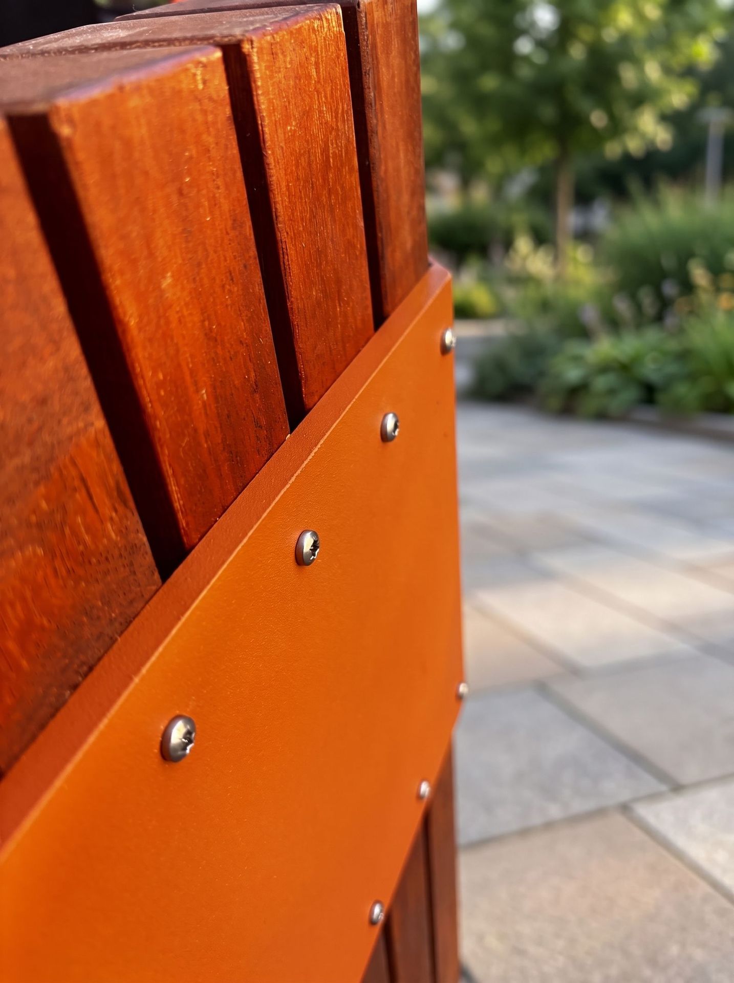 Mise en situation (décor créé par IA) : détail d’un banc public : lames d’afzélia et platine en acier vissée, sur une place pavée
